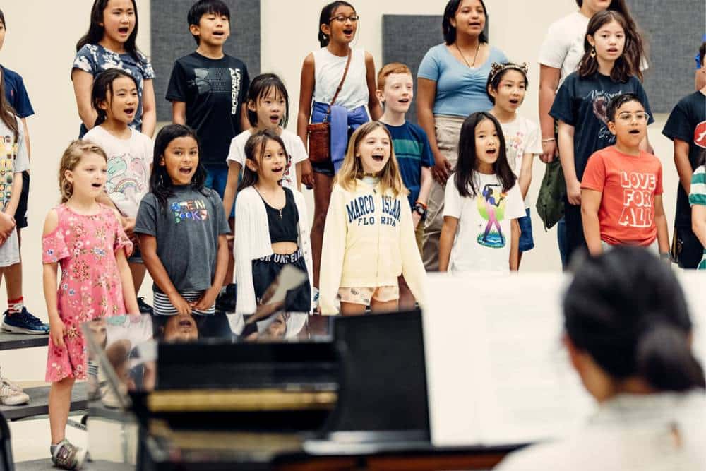 Choir students sing together as a group as their music teacher plays the piano in the foreground.