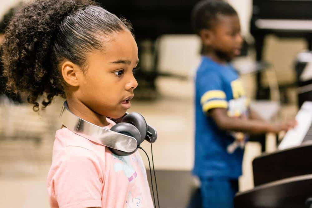 A piano student stands in front of a keyboard playing while wearing headphones. Another student plays a piano in the background.