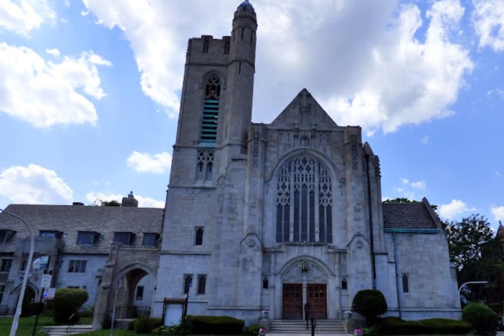The exterior of Bryn Mawr Community Church in Chicago. A grey stone church with a tall steeple and grand entrance with a blue sky and white clouds in the background.