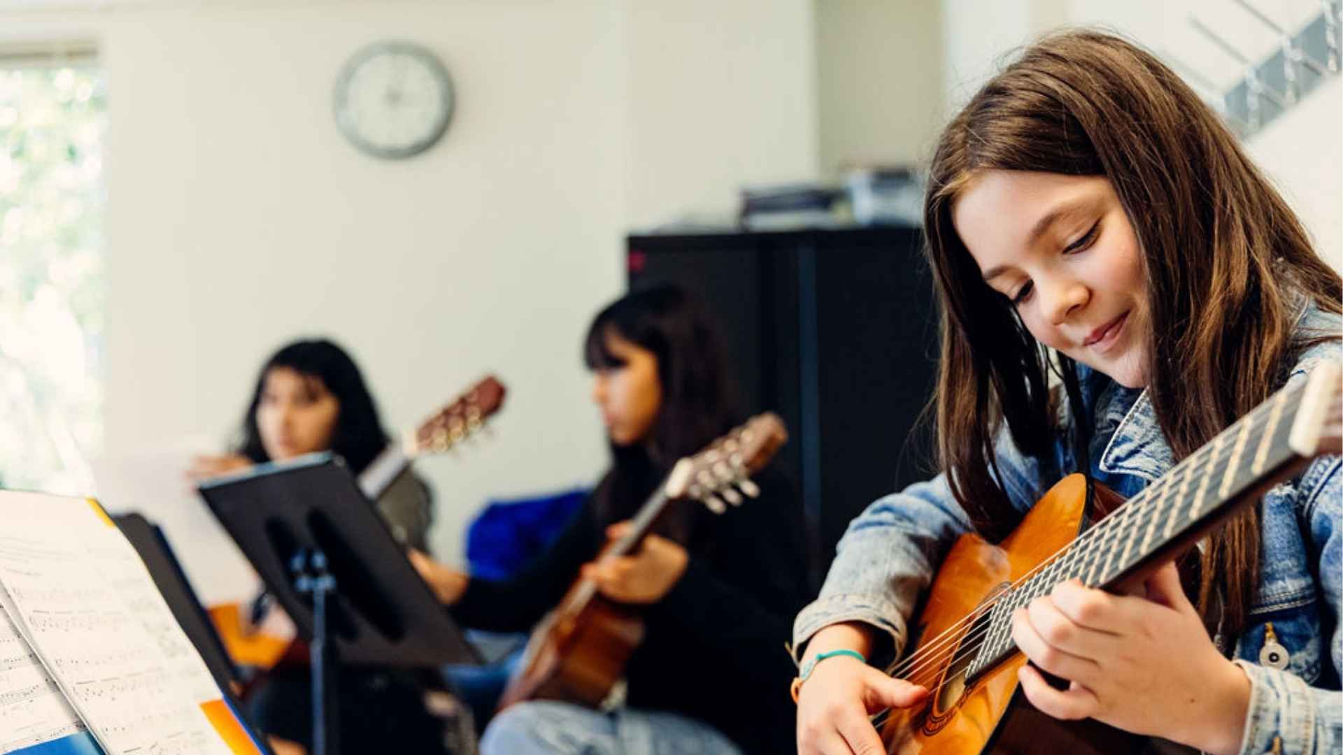 Three students in a guitar group class. Two students are out of focus in the background, while a student in the foreground plays guitar, looking down at the instrument.