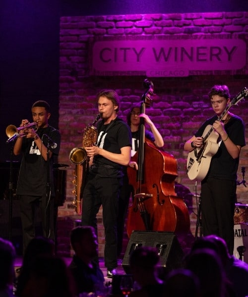 Four students playing trumpet, saxophone, double bass, and guitar perform onstage with a sign reading "City Winery" behind them