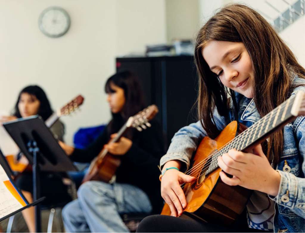 A guitar student plays their instrument while smiling. Other guitarists make music in the background.