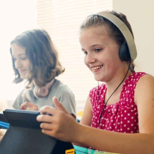 Two students are smiling, sitting next to each other and looking at their iPads. Both are wearing headphones.
