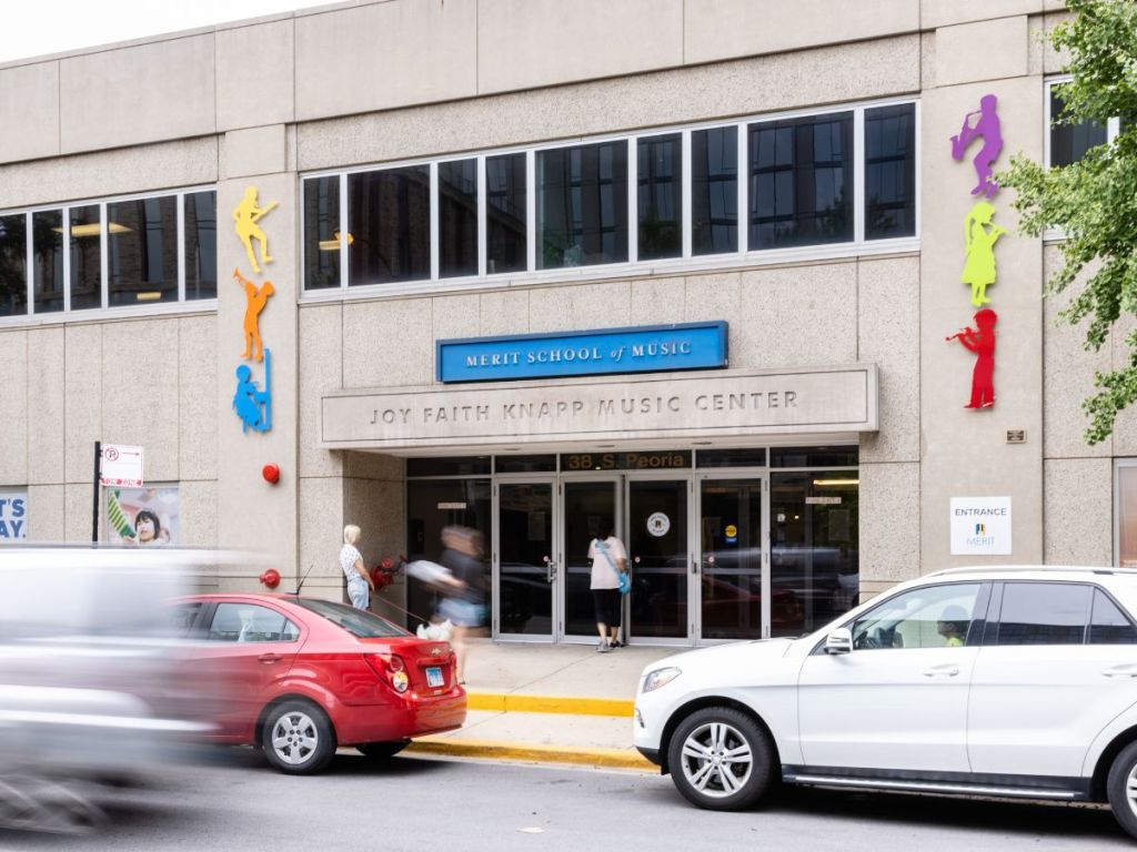 The exterior of Merit School of Music - West Loop. Cars zoom past a sand colored building with colorful sculptures of kids playing music flanking the front doors.