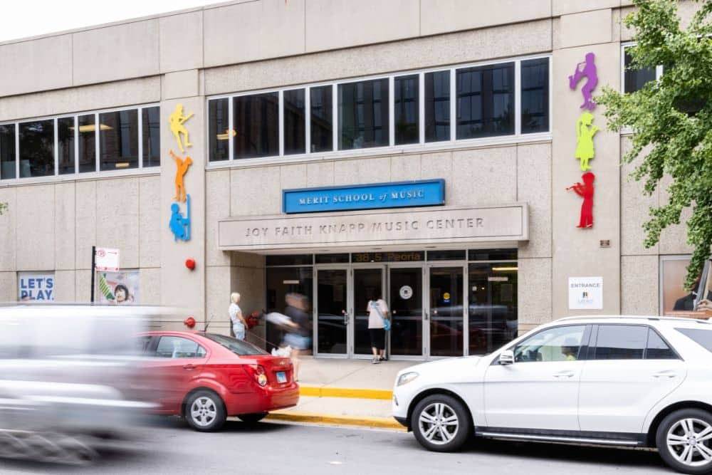 The exterior of Merit School of Music - West Loop. Cars zoom past a sand colored building with colorful sculptures of kids playing music flanking the front doors.