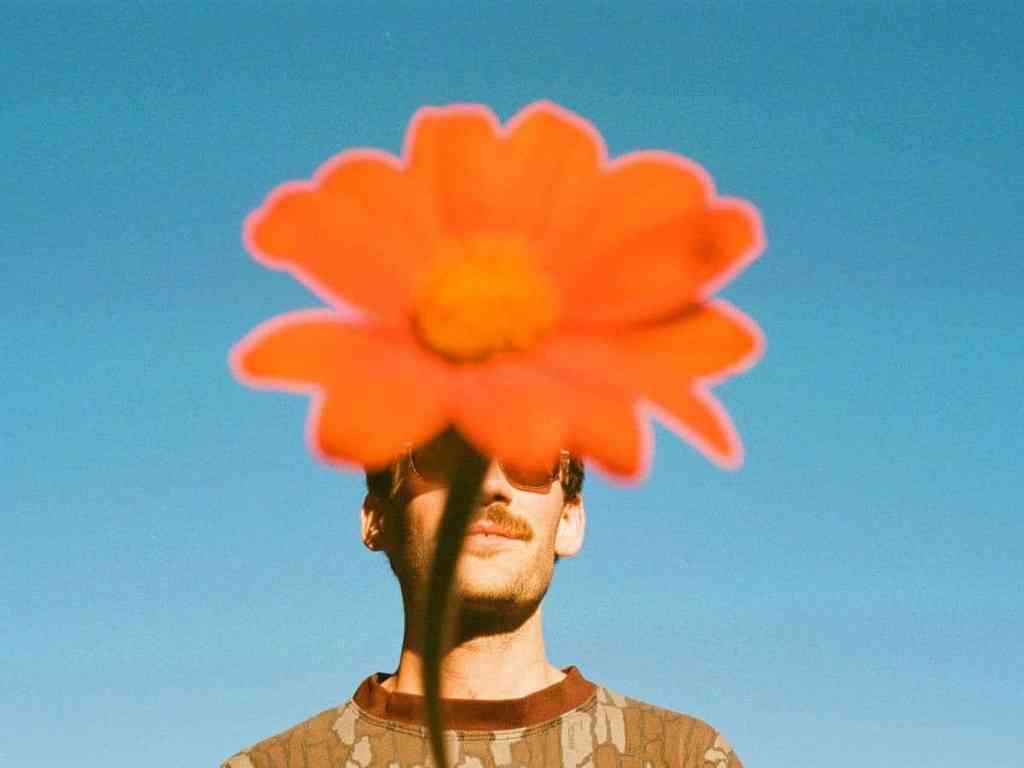 A man against a blue sky. His face is covered with a large orange flower.
