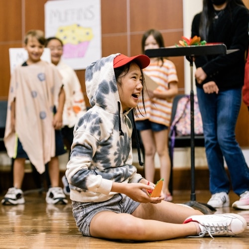 A summer camp student sits on the grown and laughs with a paper taco in hand. Other students watch in the background