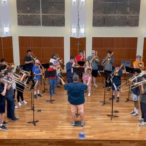 A group of students play trombone on a stage with music stands in front of them. Faculty member Timothy Riordan conducts in front of them.