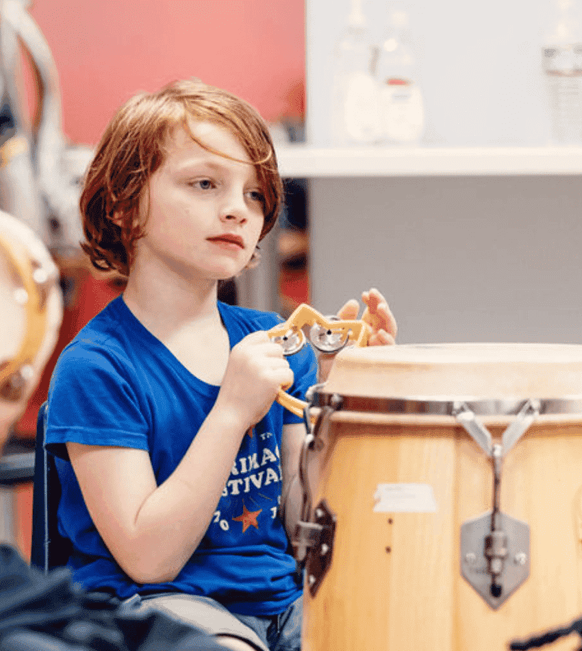 A young student holds a tambourine, surrounded by several other percussion instruments.