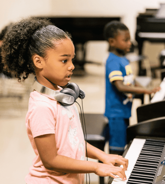 A young student plays keyboard standing up with a pair of headphones around her neck. Another student is blurred in the background.