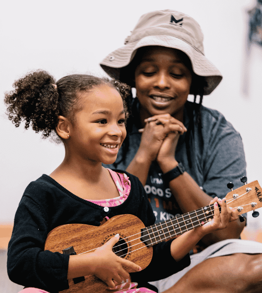 A young student plays ukulele with her parent smiling in the background.