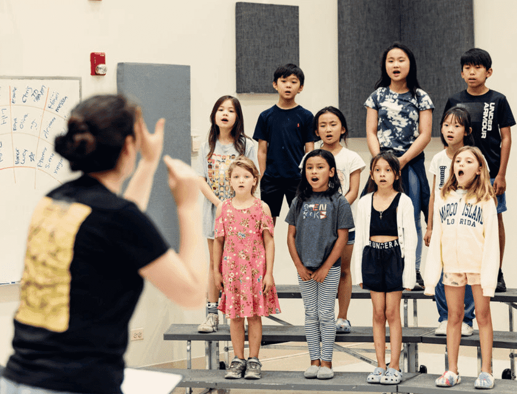 A choir of children sing together, standing on risers. In front of them is the ensemble director conducting.