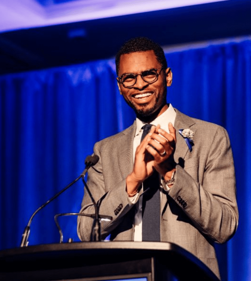 Julian Reid stands at a podium onstage in front of blue curtains, smiling and clapping.