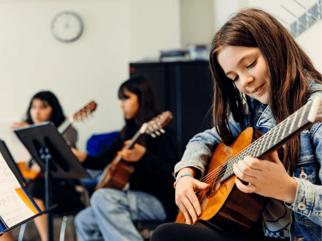 A student playing guitar in the foreground with two guitarists in the background.