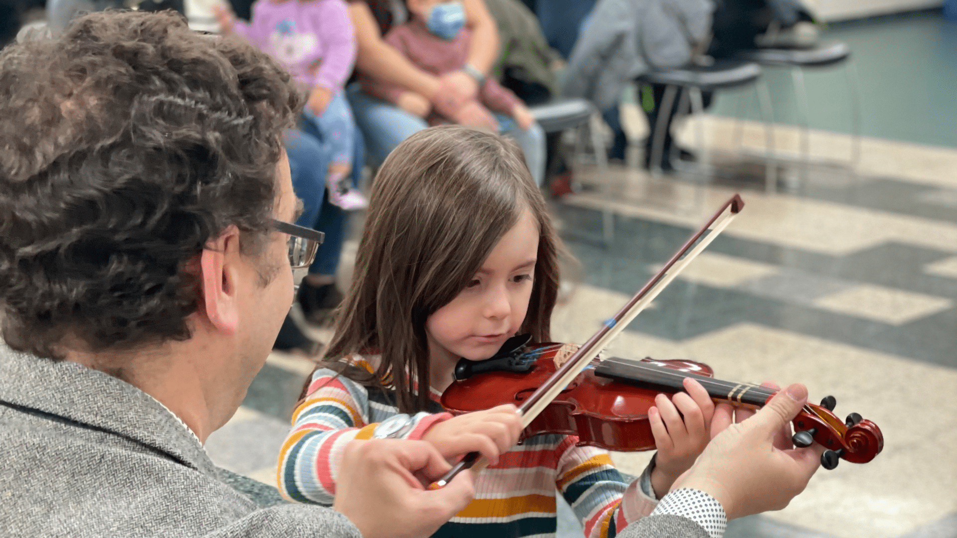 A young child holds a violin while a teacher stands in front of her, showing her how to hold it properly.