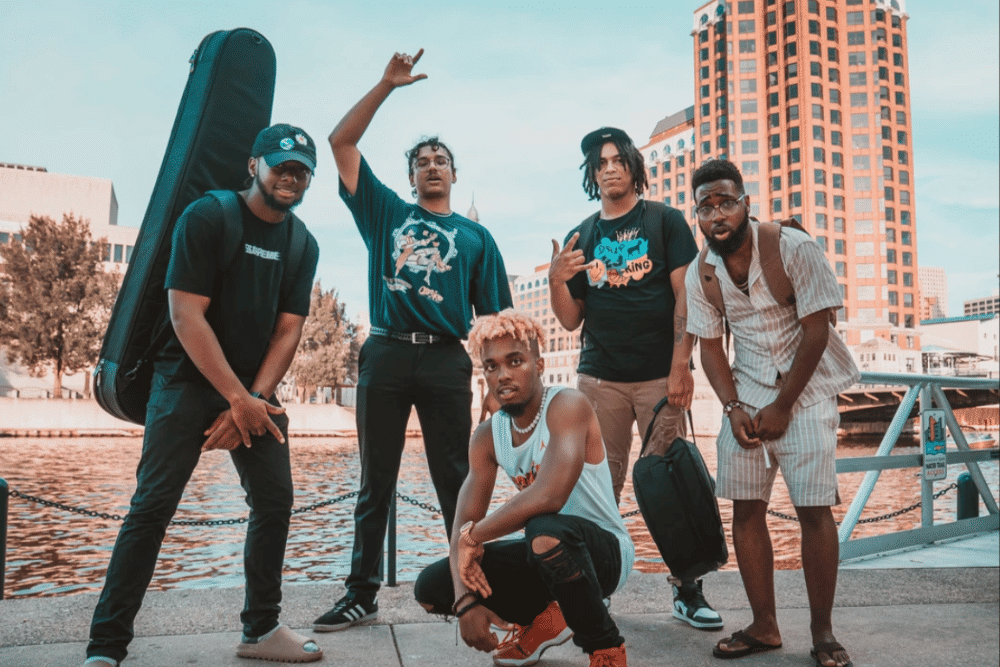 The four band members stand behind King Melik, who is crouching near the ground. They are outdoors in front of a body of water with a cityscape behind them.