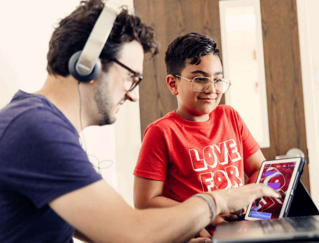 A student stands next to Douglas Orofino, who is seated at a piano. Orofino is reviewing the student's piece on his iPad.