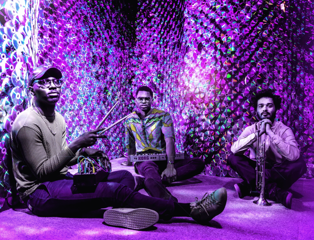 Nova Zaii, Julian Reid, and Nico Segal of The JuJu Exchange sit in a room with purple lights and CDs on the walls.