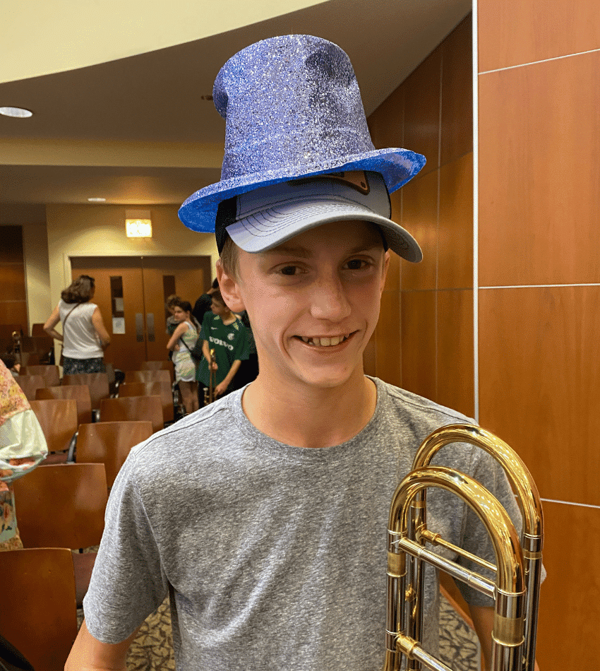 Student Braden W. poses holding his trombone. He is wearing two hats: a baseball cap, and on top of it is the Camp Hat, a sparkly purple top hat.