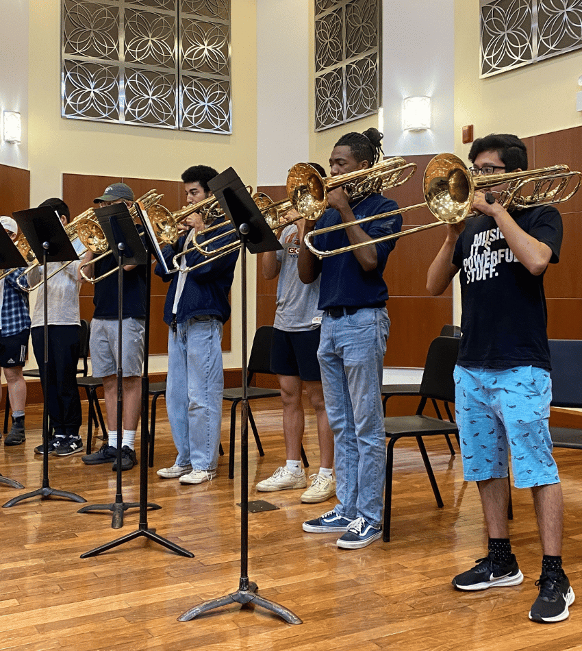 Several students playing trombone look at music stands in front of them as they rehearse onstage.