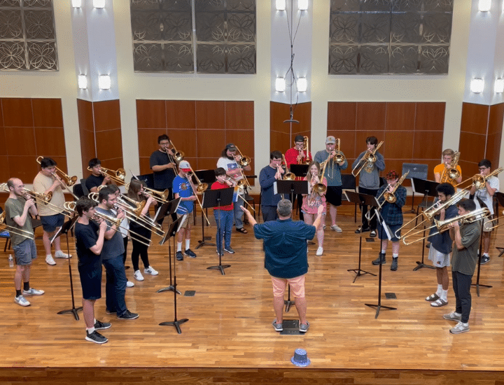 A group of students play trombone on a stage with music stands in front of them. Faculty member Timothy Riordan conducts in front of them.